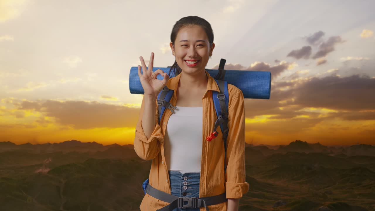 excursionista asiática con mochila de montañismo sonriendo y mostrando un gesto de acuerdo a la cámara mientras está de pie en la cima de la montaña durante la hora del atardecer