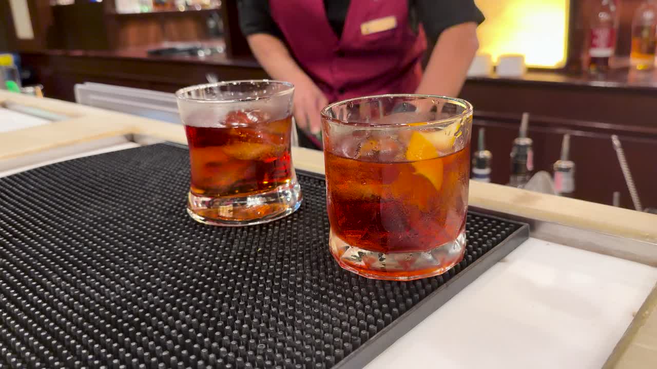Bartender in maroon uniform serves Negroni and Manhattan cocktails on bar mat, warm lighting