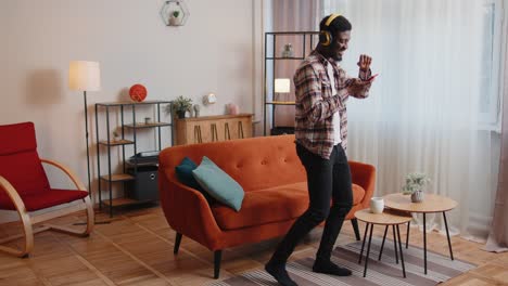 joven encantado con auriculares inalámbricos bailando, cantando en un sofá acogedor en la sala de estar en casa