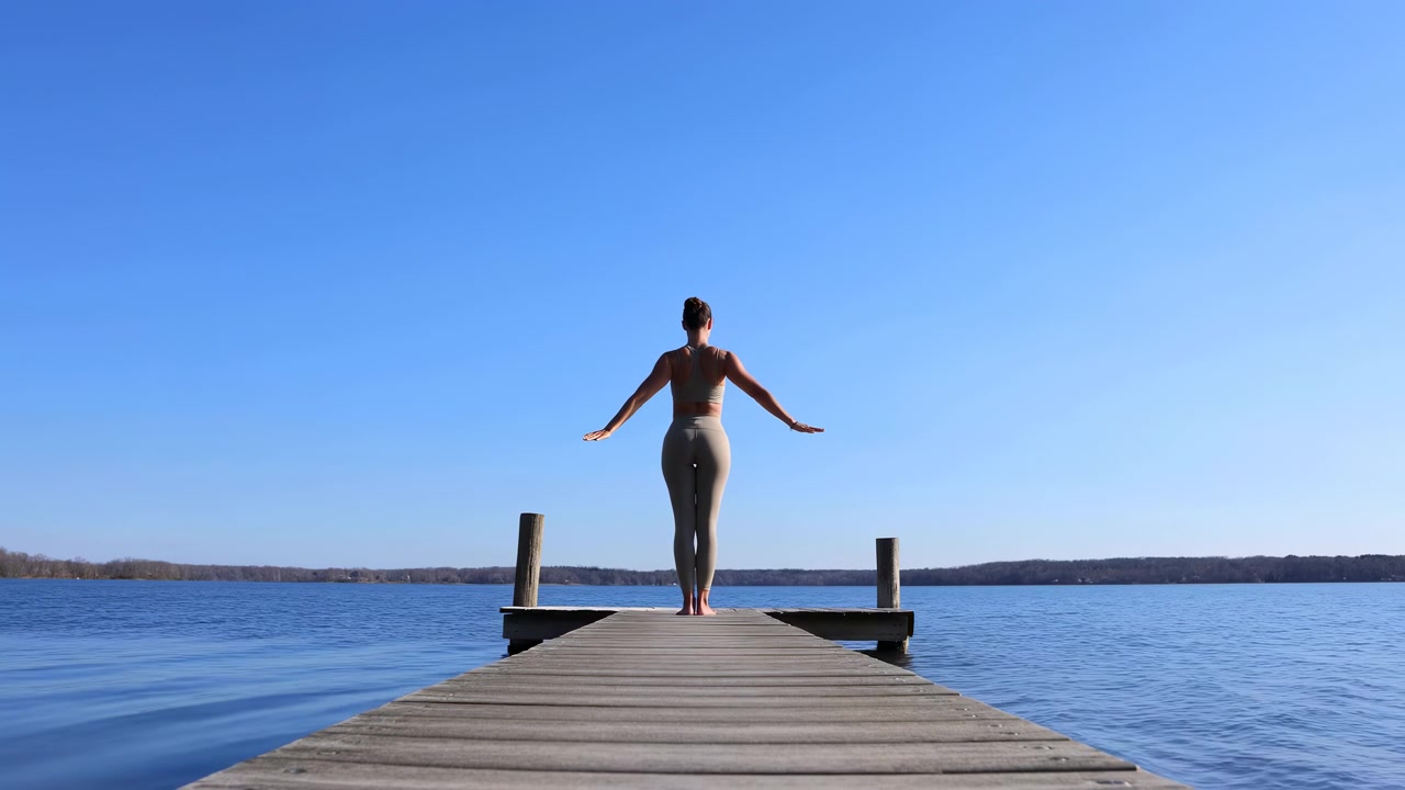 Vista posteriore di una donna in piedi all'estremità di un molo di legno, con le braccia leggermente estese, incorniciata da un cielo azzurro limpido e una riva alberata in lontananza. Luce diurna soffusa.