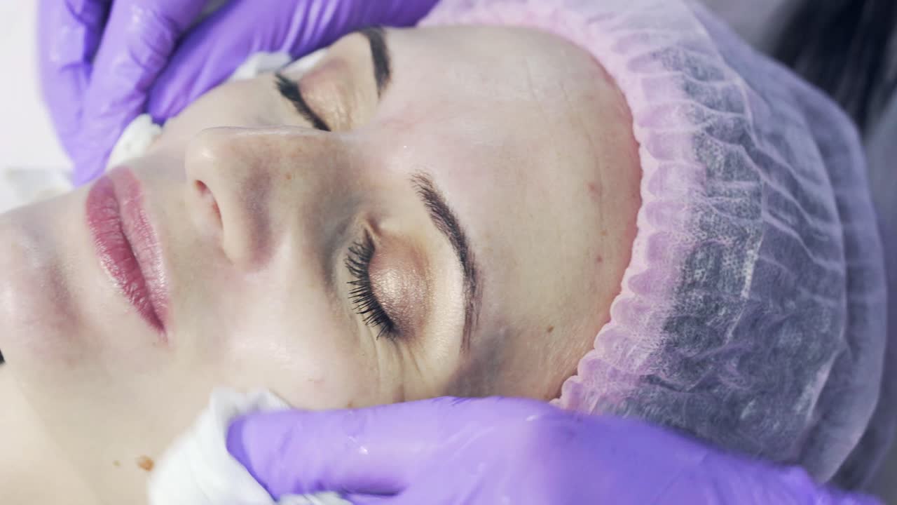 Close up of hands of skillful beautician cleaning and touching female face with napkin in Spa clinic. The woman is lying and relaxing.