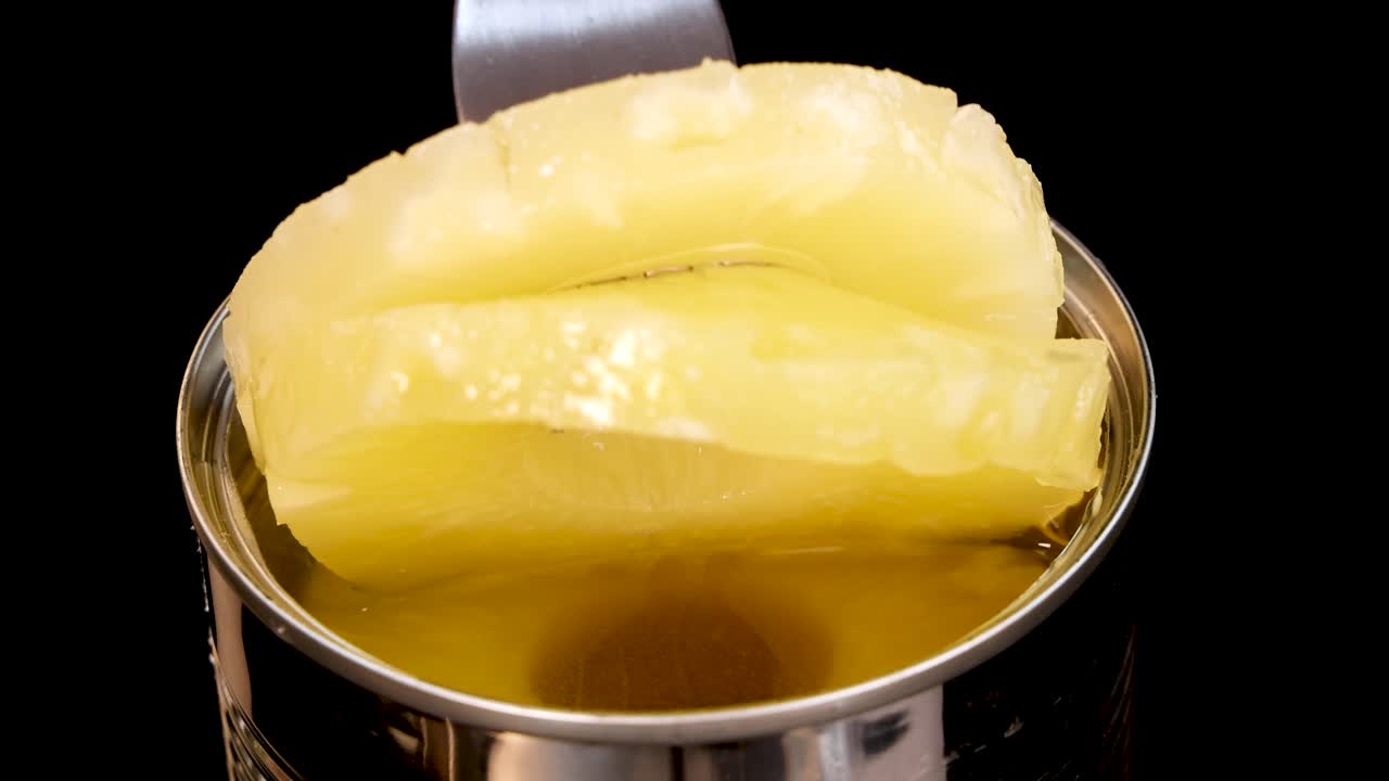 A metal fork lifts a pineapple ring from an opened can, with bright studio lighting and a clean, isolated black background. Smooth, overhead camera angle