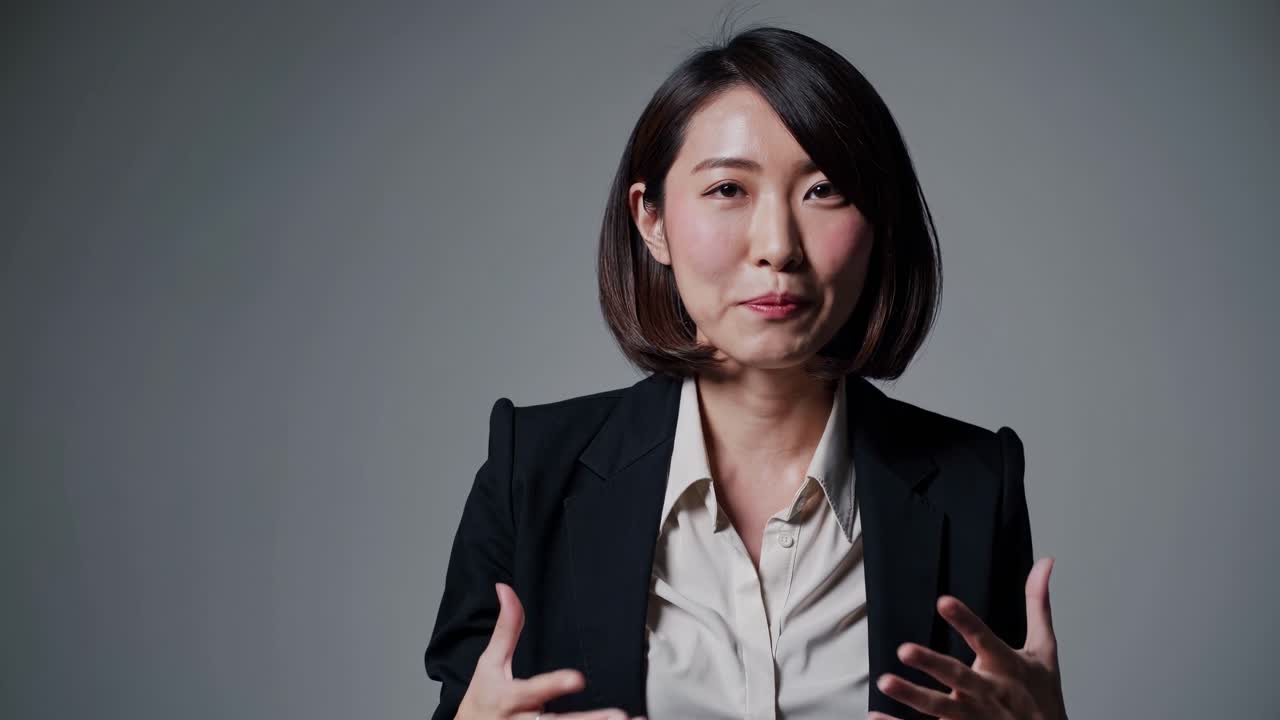 A professional woman in a suit speaks directly to the camera in a video, using a medium close-up