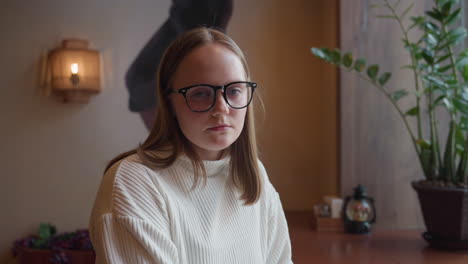 student sits beside large window in cozy cafe, looking serious while holding mug, indoor plant placed near her on wooden table, wall art and snowy cityscape seen outside create serene background