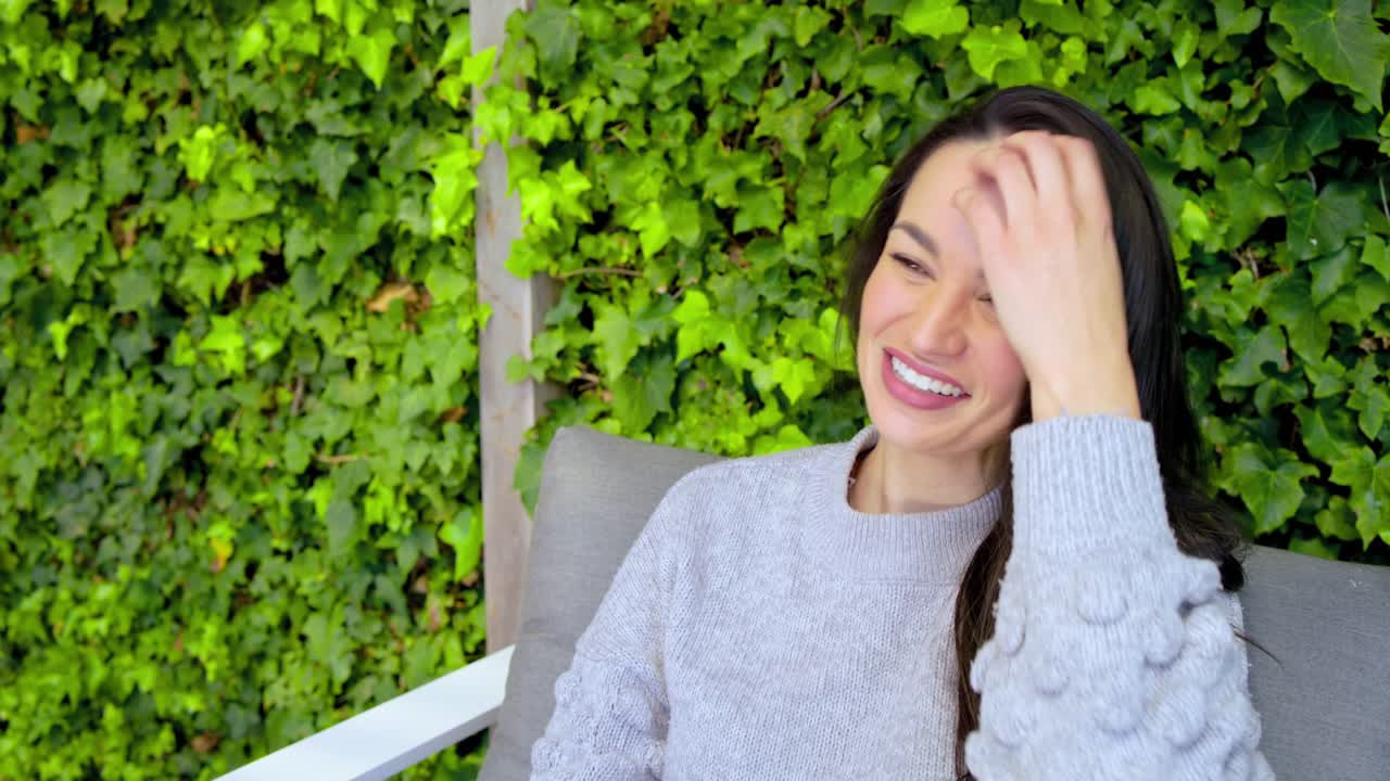 Mid adult woman sitting on white-framed lounge chair on patio under ivy wall laughing brushing hair