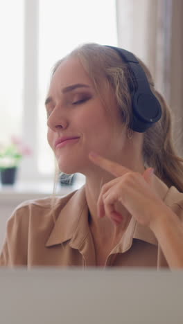 young blonde woman in beige shirt moves to music beat wearing black headphones at break against bright window in home office slow motion closeup