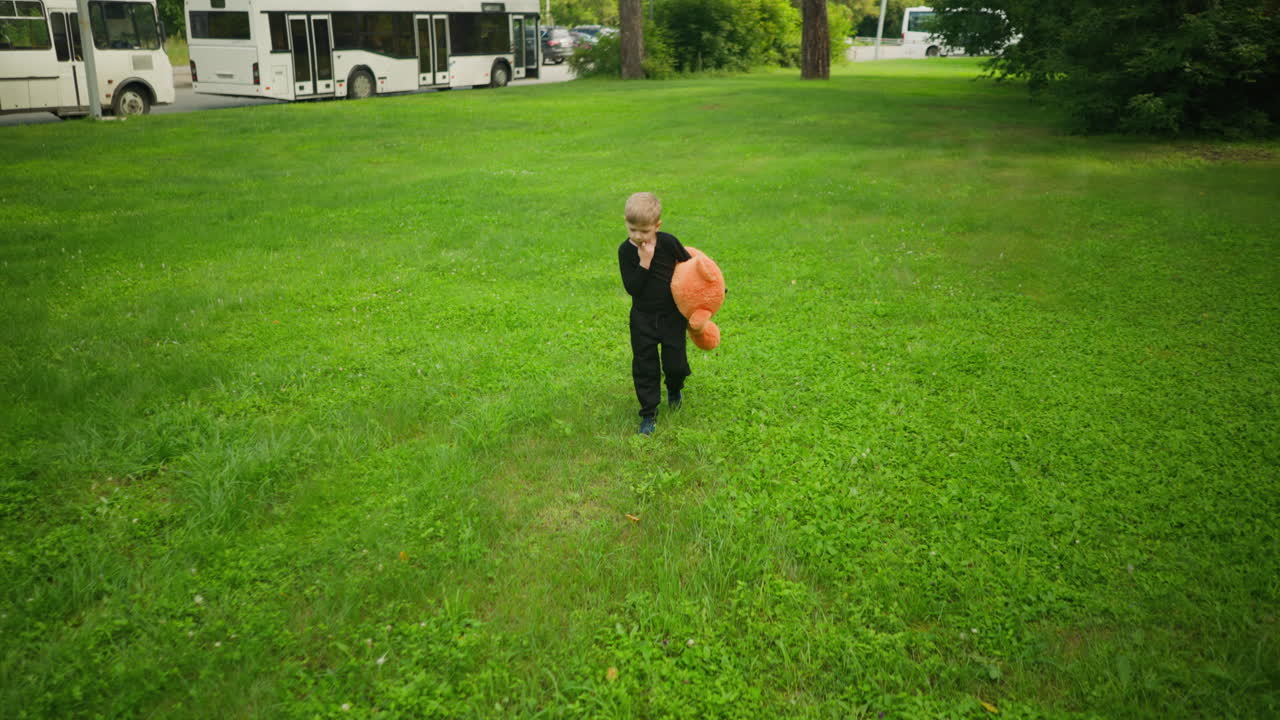 Little child in black outfit walking across lush green grassy field holding large orange teddy bear, with several white buses parked in background under natural daylight surrounded by trees
