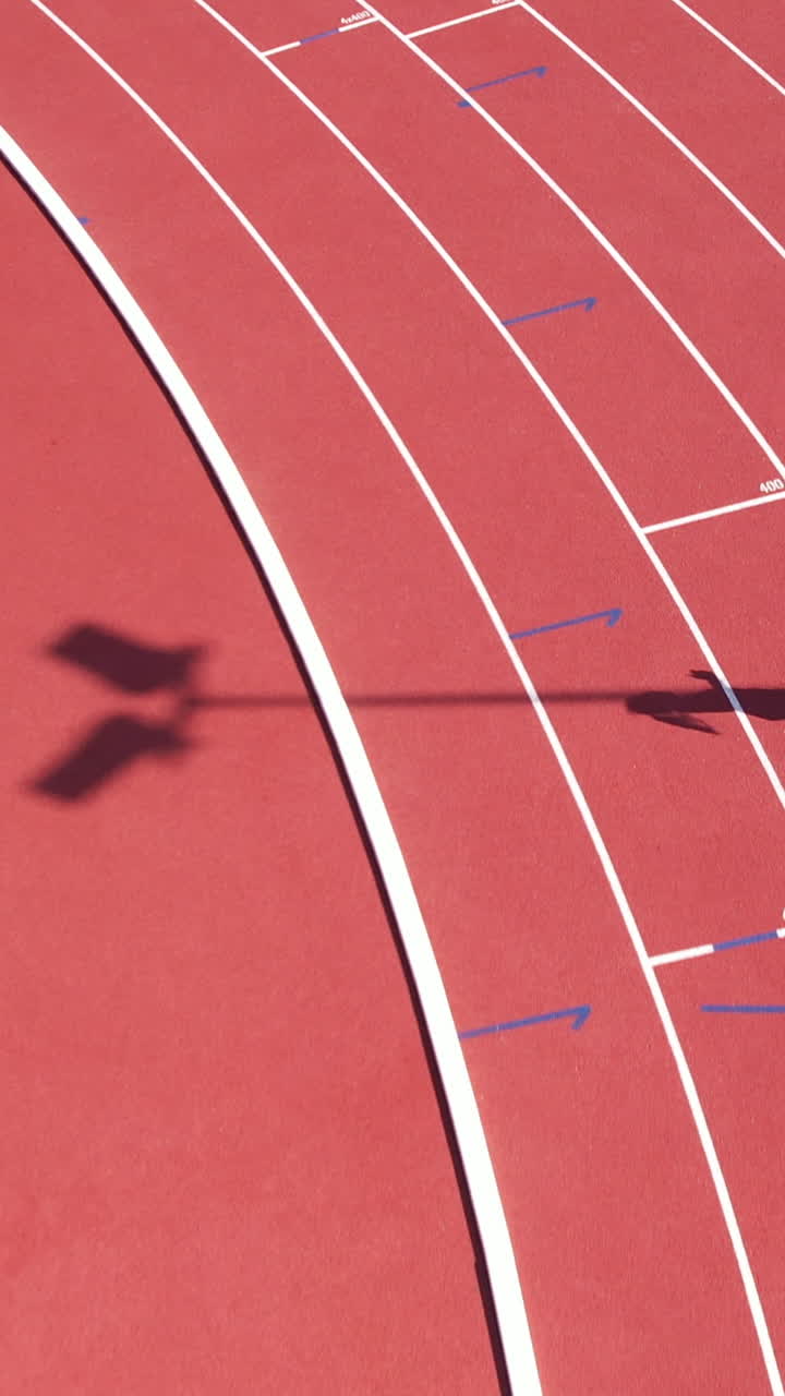 Inquadratura verticale dall'alto dell'ombra di un'atleta che corre su una pista rossa in uno stadio sportivo. Ideale per contenuti di fitness e motivazionali.