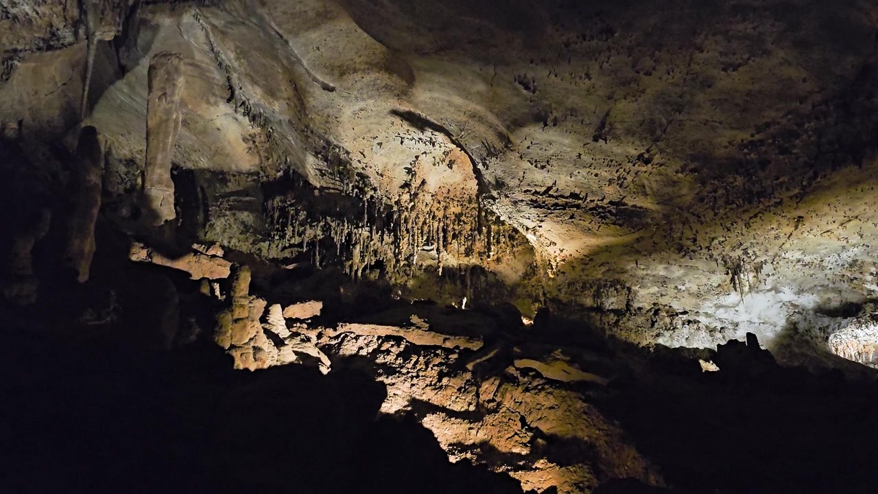 Dramatic cave landscape with jagged stalactites hanging above rocky formations, illuminated limestone textures, deep shadows, and natural underground geology details.