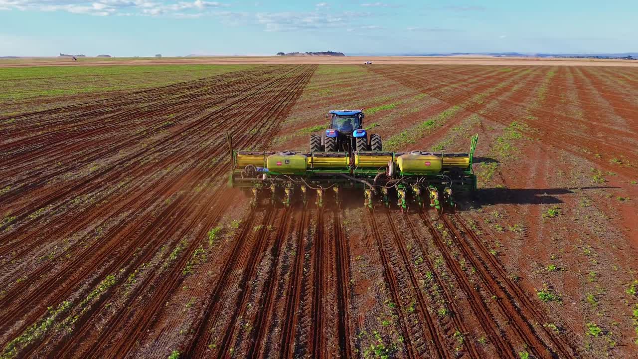 Trattore blu che traina una seminatrice di precisione a più file a Formosa, Goiás, Brasile centro-occidentale, creando file di semina uniformi con spaziatura precisa nell'agricoltura meccanizzata, ripresa con drone in avvicinamento.