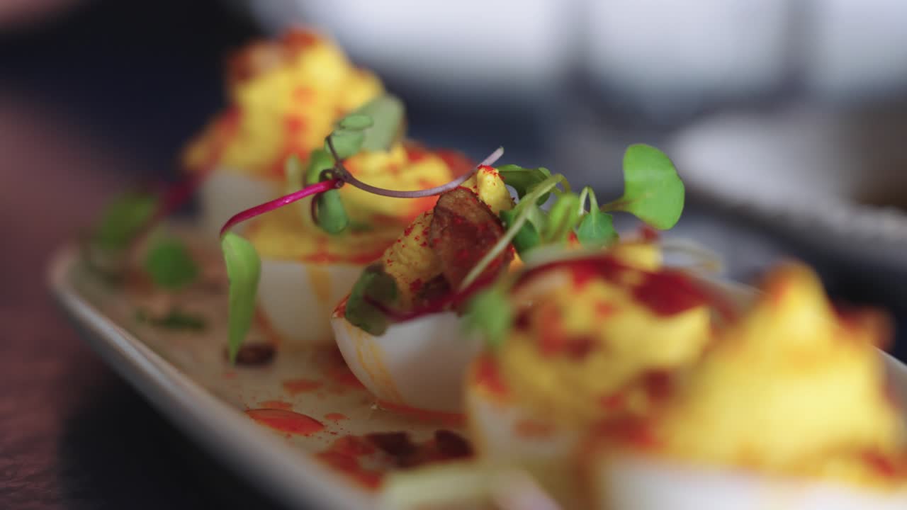 Filmato 4K in slow motion con avvicinamento di uova alla diavola irrorate con salsa e guarnite con pancetta e microgreens, in un elegante ristorante per il brunch, che mette in risalto la presentazione gourmet dell'antipasto.