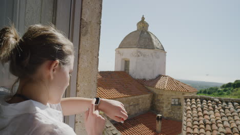 mujer usando reloj inteligente comprobando mensajes disfrutando de un cálido día soleado de vacaciones de pie en el balcón turista feliz relajándose en italia