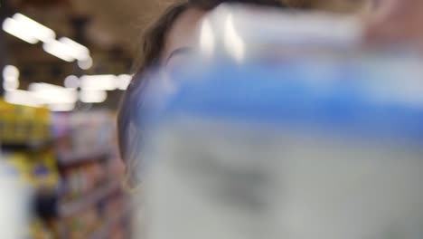 Close-up-portrait-of-a-young-curly-haired-woman-doing-grocery-shopping-at-the-supermarket,-she-is-reading-a-product-label-and-nutrition-facts-on-a-box.-Footage-from-the-shelf