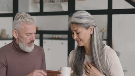 middle aged woman having chest pain and her husband giving her a hot drink while they sitting together on sofa at home