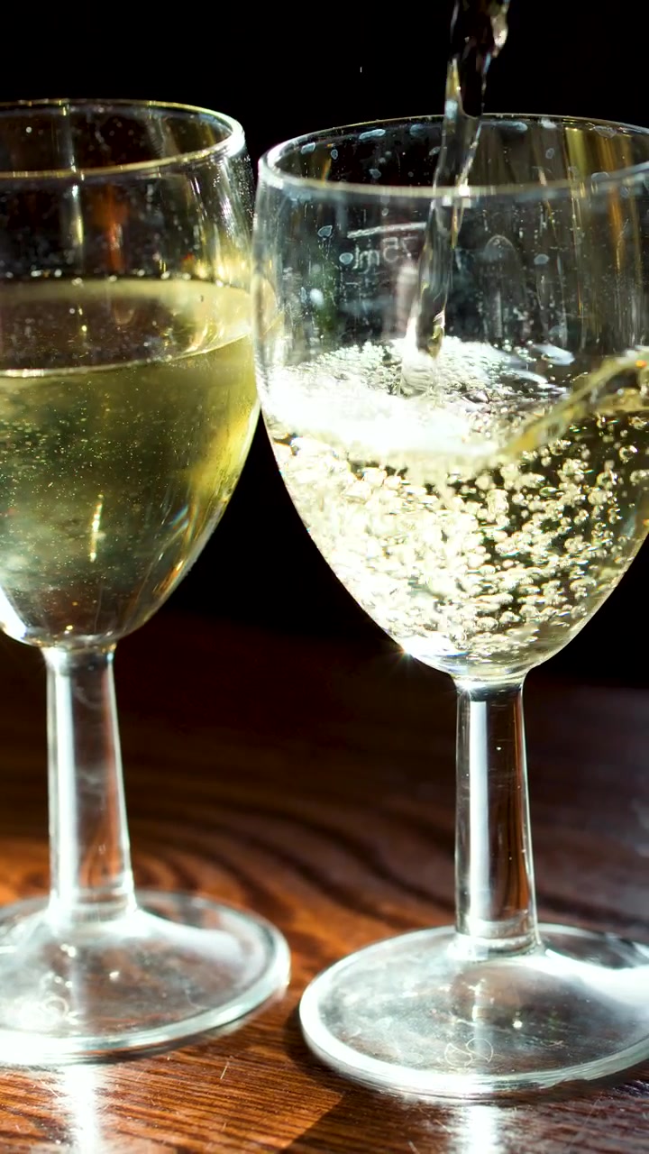 Vino bianco frizzante versato in bicchieri, illuminazione laterale, primo piano, superficie in legno, effetto slow-motion.