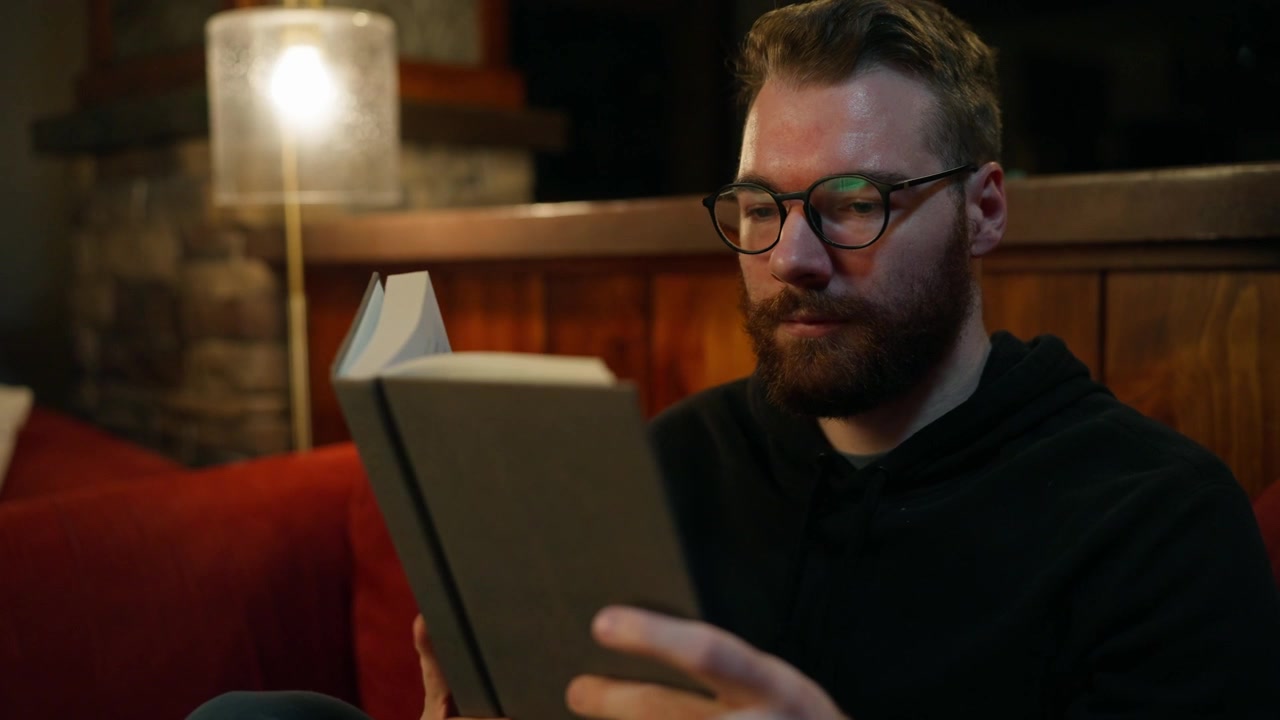 Man with glasses reading a book indoors on a cozy sofa, with warm lamp light, wooden interior and calm evening lifestyle atmosphere.