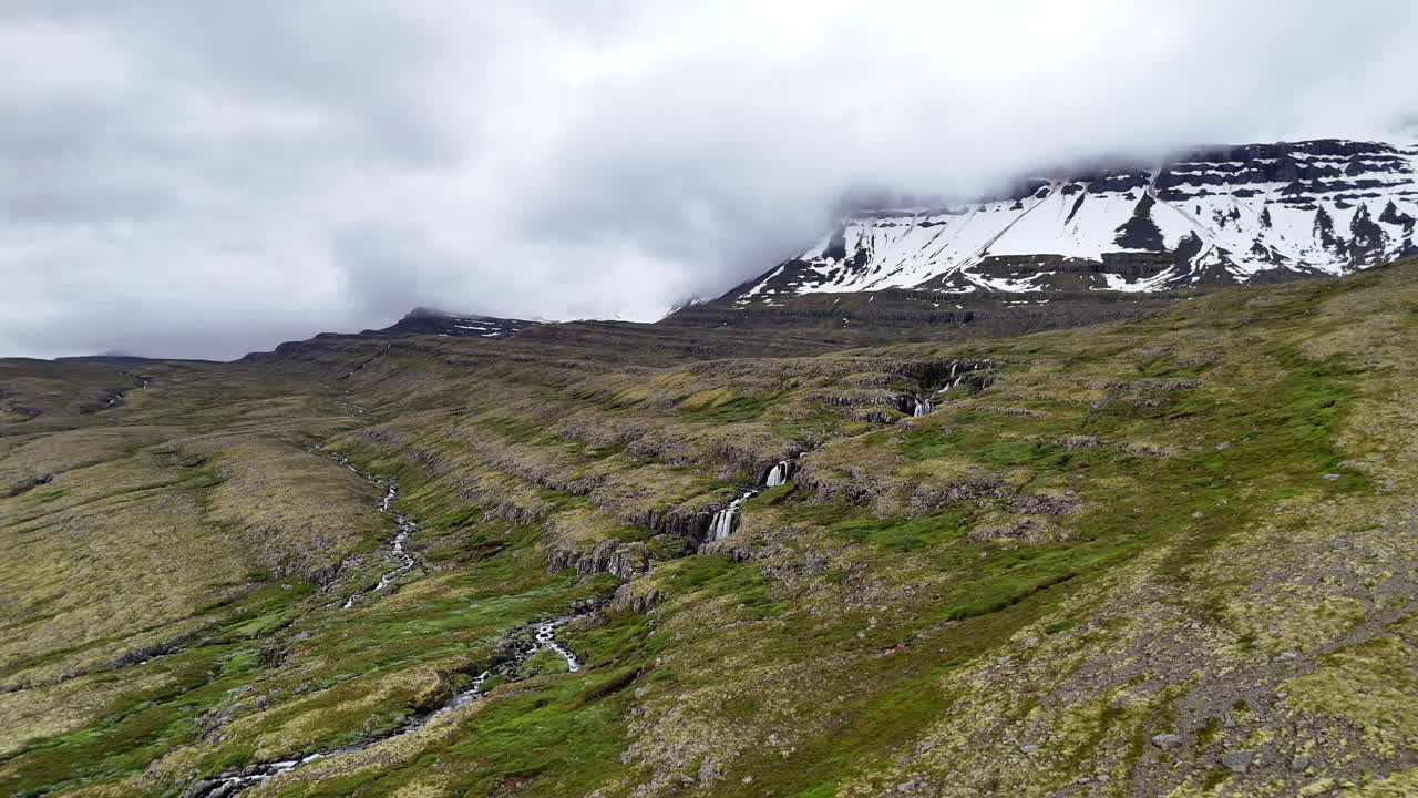 Wide aerial view of the mountains near Fáskrúðsfjörður, with snow-lined peaks, scattered waterfalls, winding wetlands, and an open valley crossed by a curving road under cloudy skies