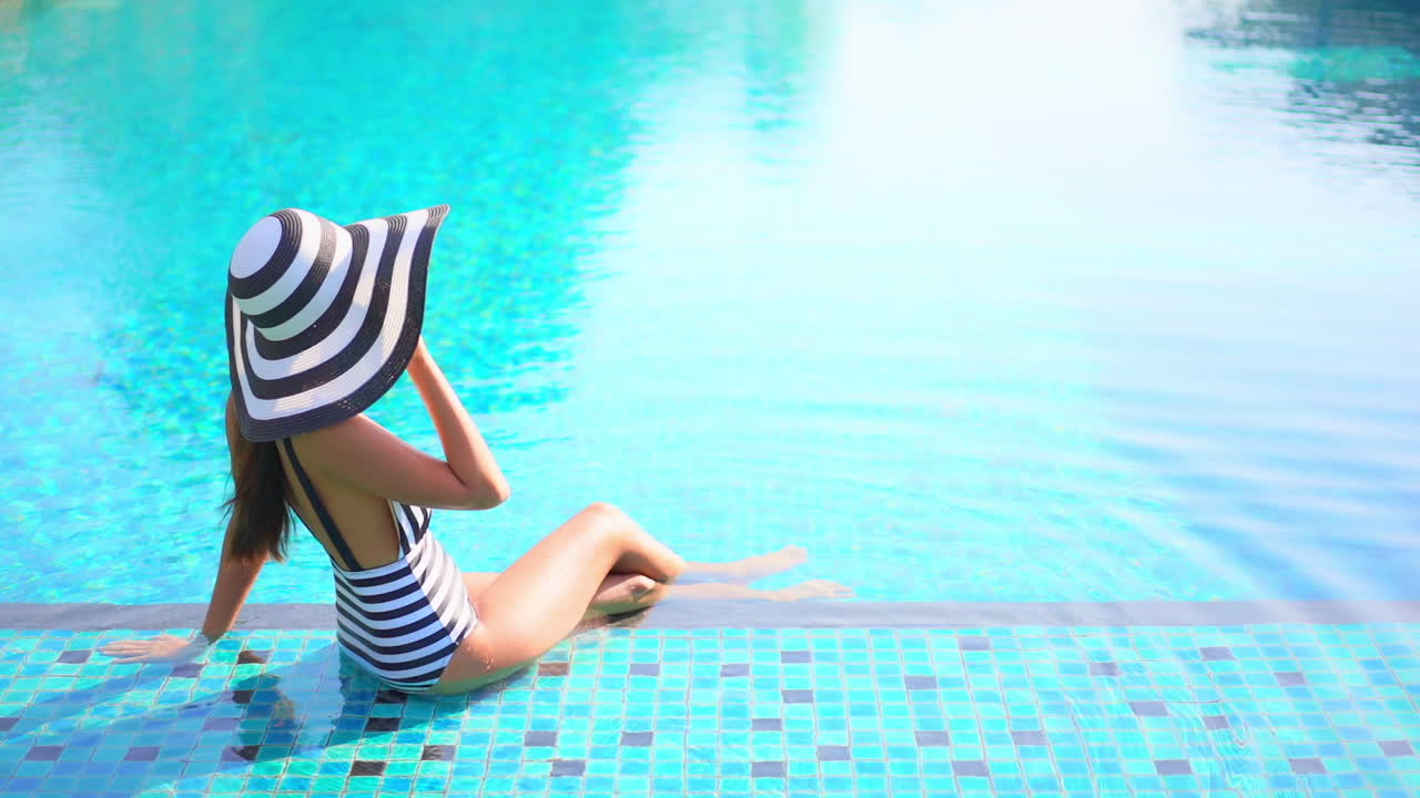 Back to the camera A young woman sits in the shallow end of a resort swimming pool, grabs the brim of her black and white sun hat as the wind blows
