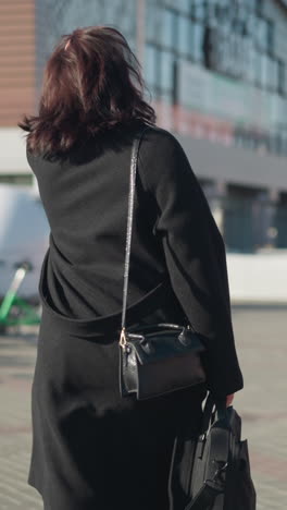 vista trasera de una mujer con abrigo negro y bolso de mano caminando por una calle urbana soleada cerca de bicicletas verdes, con arquitectura moderna y fondo borroso