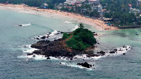 tropical sri lanka aerial view of a rocky islet covered in lush greenery, surrounded by turquoise waters and crashing waves, near a sandy beach under a bright, sunny sky, captured with wide-angle lens