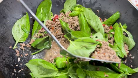 minced meat, fresh basil leaves, garlic, and chopped chilies are stir-fried in a wok under bright lighting, with visible spoon movement and sizzling action