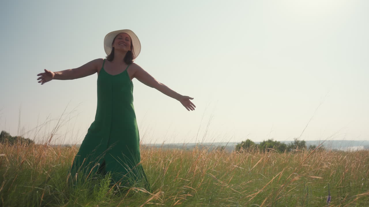 happy woman in green dress and white hat smiling with arms wide open dancing joyfully in sunlit grassy field during summer adventure with distant parked car and trees under bright clear sky