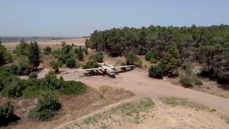 aerial slow forward, zoom in drone shot of an old dismantled army plane monument placed on an empty dirt field, surrounded by trees and bushes