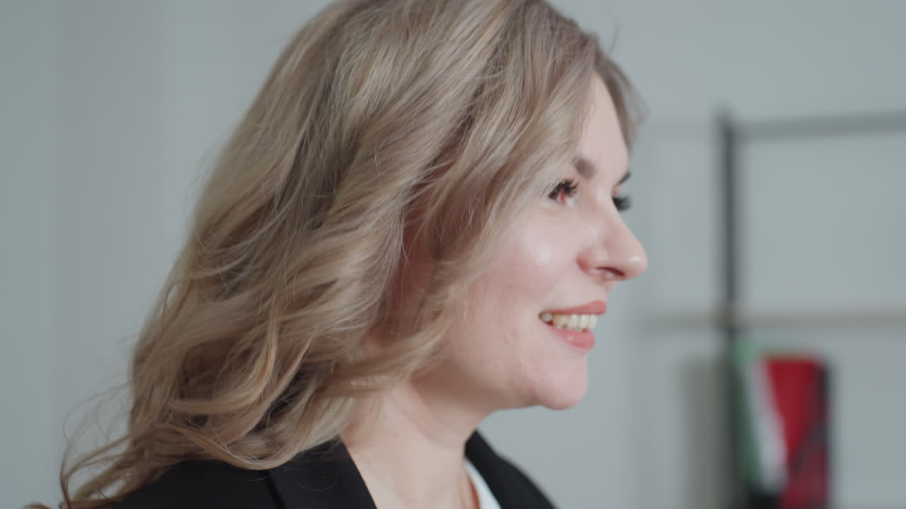 Close up side view of cheerful woman smiling as she walks into office, background shows blurred shelf and wall with colorful sticky notes creating casual creative workplace atmosphere