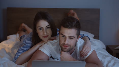 young caucasian couple lying in the night on the bed and watching a video on the laptop