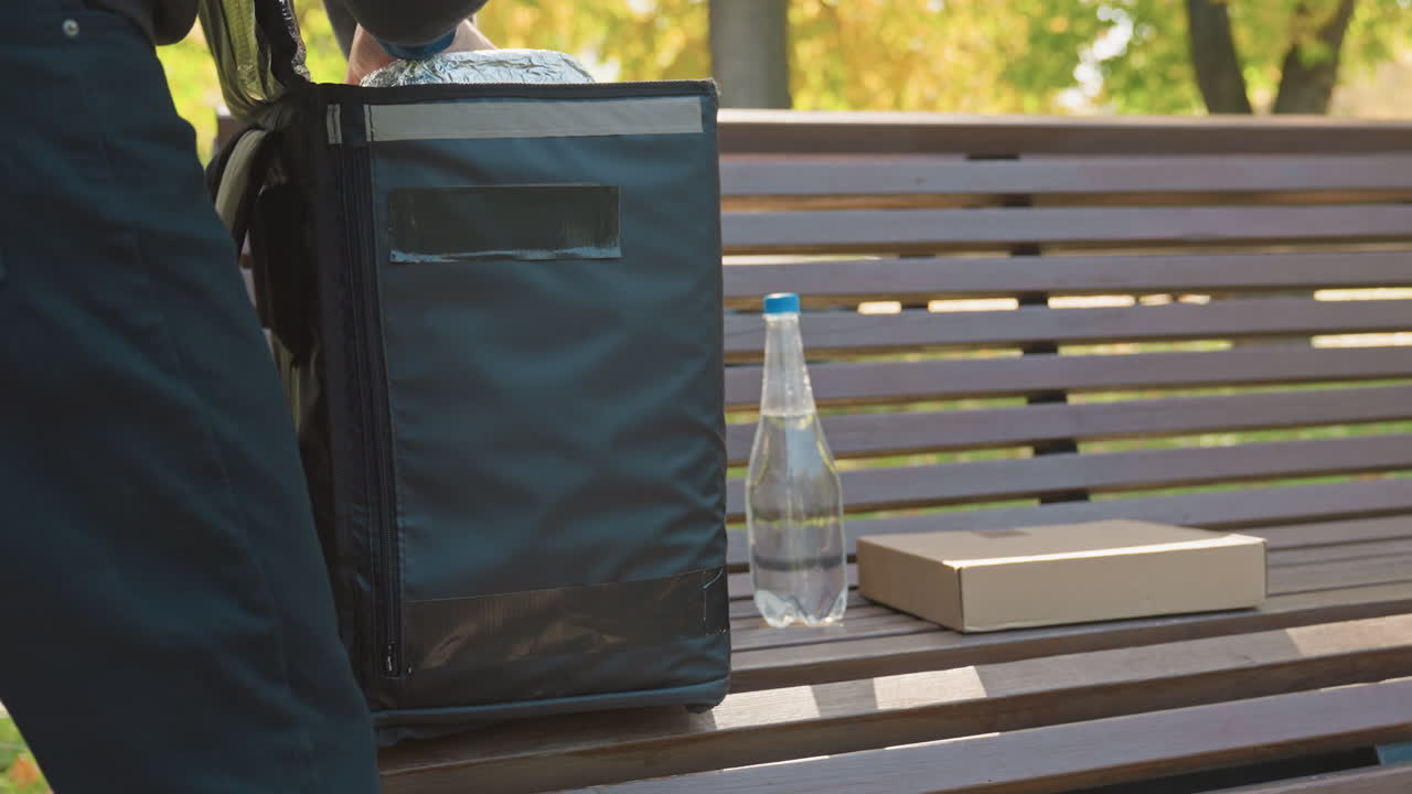 entrega de comida sin contacto en un banco, mochila isotérmica junto a una botella de agua y una caja de cartón, mensajero cogiendo el pedido en un parque frondoso, listones de madera, luz cálida de otoño, uniforme informal y ambiente urbano tranquilo