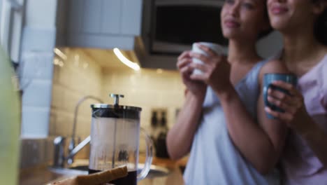 Mixed-race-lesbian-couple-hugging-and-drinking-coffee-in-kitchen