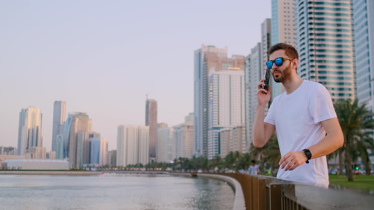 A young male tourist in sunglasses and bristles hipster in a white t-shirt standing on the waterfront against the background of the modern city writes text messages phone for social networks is live broadcast