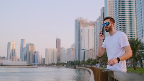 Ein-Junger-Männlicher-Tourist-Mit-Sonnenbrille-Und-Borsten,-Ein-Hipster-In-Einem-Weißen-T-Shirt,-Der-An-Der-Uferpromenade-Vor-Dem-Hintergrund-Der-Modernen-Stadt-Steht,-Schreibt-Textnachrichten,-Das-Telefon-Für-Soziale-Netzwerke-Wird-Live-übertragen