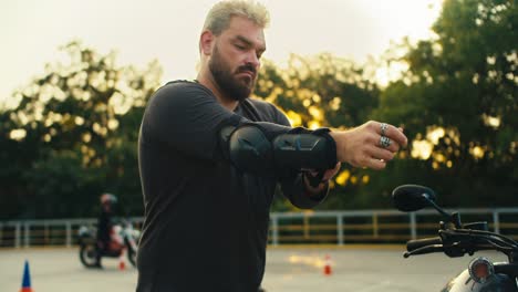 a man biker and strong build with a thick beard puts on elbow pads for protection while eating on a motorcycle