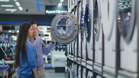 a handsome man and a woman choose a washing machine to buy open the door and inspect the coating of the drum and the quality of the product. woman looking inside washing machine