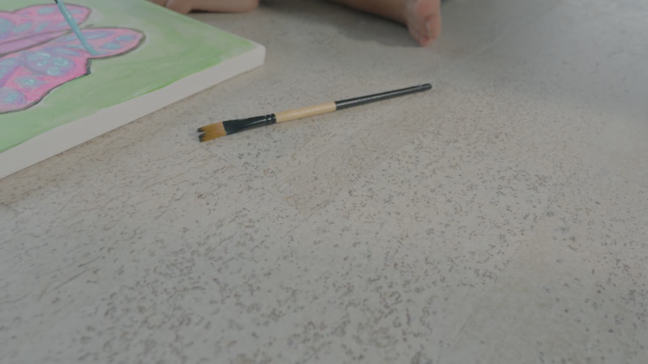 Low tracking shot of a young girl sitting on the floor painting a colourful butterfly on a canvas