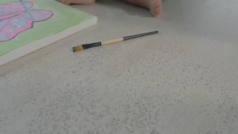 low tracking shot of a young girl sitting on the floor painting a colourful butterfly on a canvas