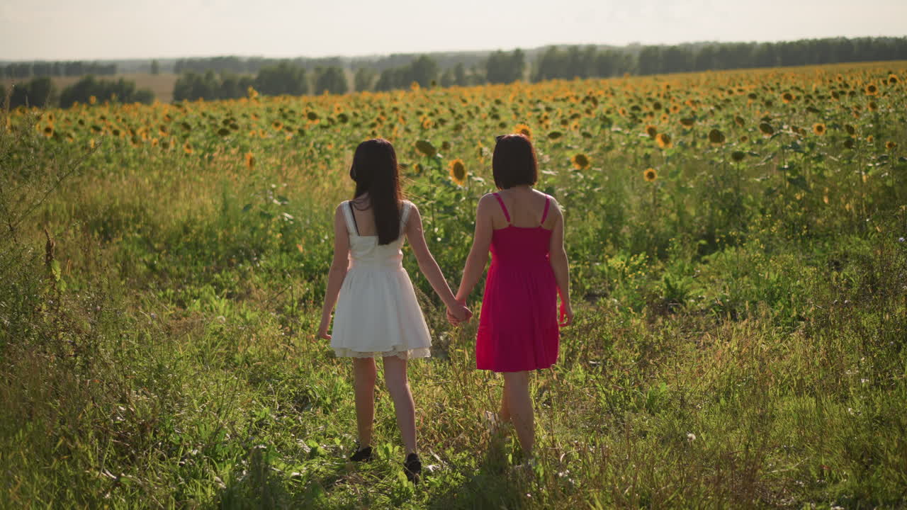 Dos mujeres caminando hacia un campo de girasoles, vista trasera de una pareja tomándose de las manos, infinitos girasoles hasta el horizonte, cálida luz de tarde, sendero rústico en el campo, vestidos informales y botas, amplia panorámica cinematográfica
