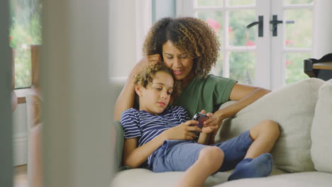 Madre-E-Hijo-Sentados-En-El-Sofá-De-Casa-Jugando-Juntos-A-Videojuegos-En-El-Teléfono-Móvil