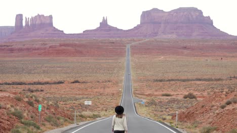 mujer joven con sombrero caminando en la famosa carretera vacía que conduce a las colinas de arenisca del parque tribal navajo del valle del monumento en la frontera de arizona-utah en estados unidos - cámara lenta inclinándose hacia arriba