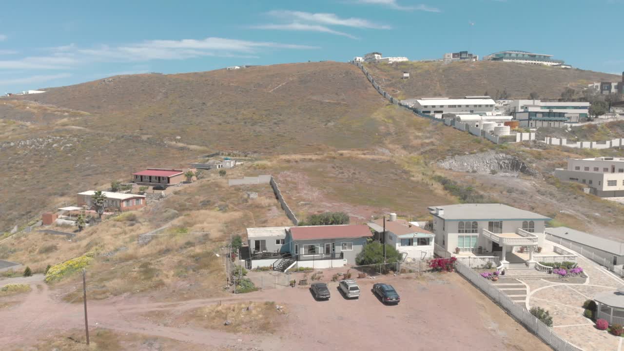 Ripresa aerea con drone che si allontana di una casa in collina con balcone e case circostanti, terreno arido e auto parcheggiate visibili, ampio paesaggio che rivela un contesto e un ambiente suburbano.