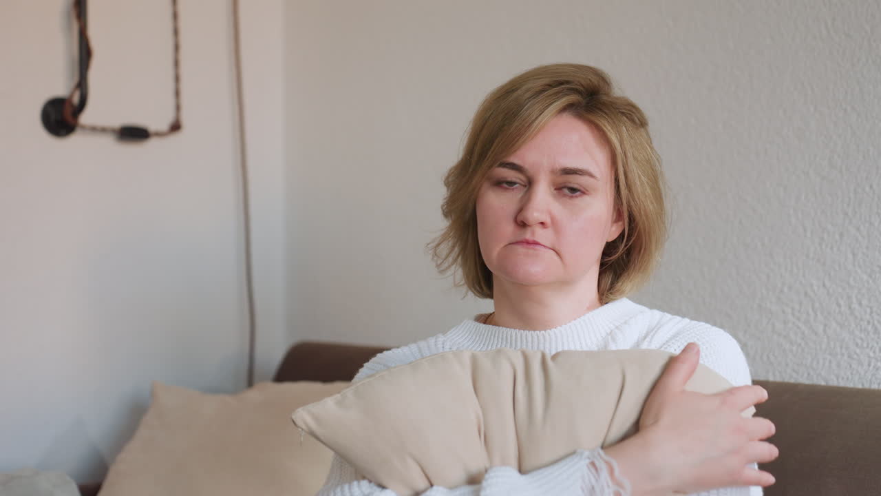 Portrait of female client sitting on brown sofa holding pillow close, wearing white sweater, reflecting quietly in calm therapy setting with soft lighting and neutral interior background