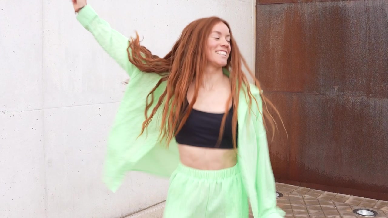 A youthful lady is seen dancing energetically in a bright green attire outdoors under the sunlight