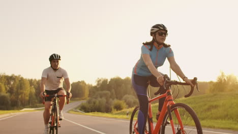 Un-Hombre-Y-Una-Mujer-En-Bicicleta-Recorren-Juntos-La-Carretera-Al-Atardecer-En-Cámara-Lenta.-La-Pareja-Viaja-En-Bicicleta.-Cascos-De-Ciclismo-Deportivos.