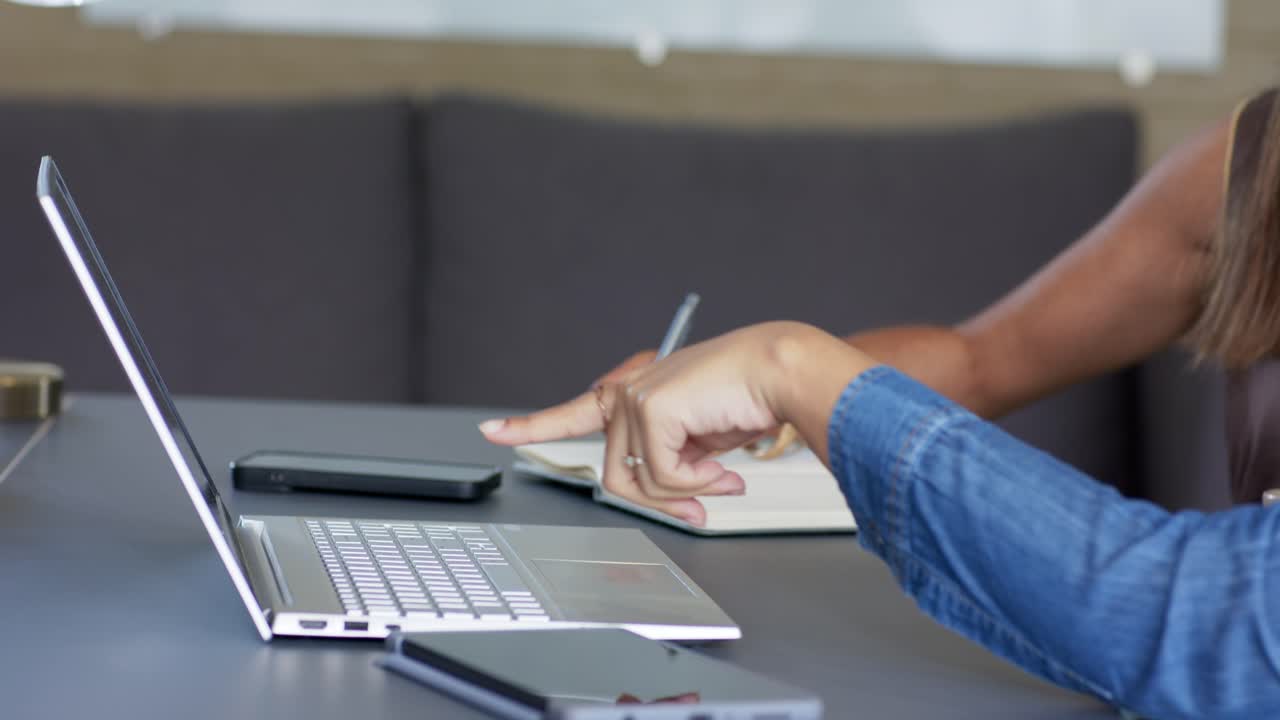 Una mano entra en el encuadre, navegando por un portátil desplazándose por el trackpad y, a continuación, tomando notas en un cuaderno.