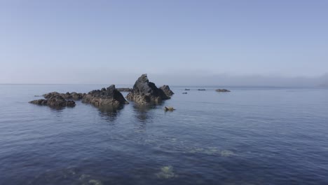 low aerial skims water surface toward rugged eroded sea rocks in ocean