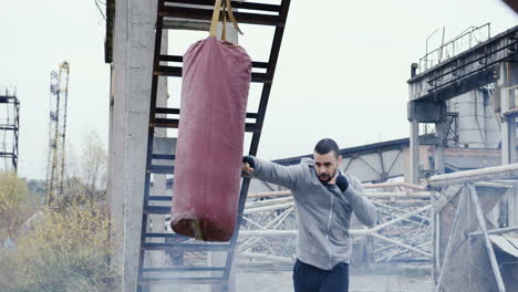 caucasian man in grey hoodie hitting a punching bag outdoors an abandoned factory on a cloudy morning