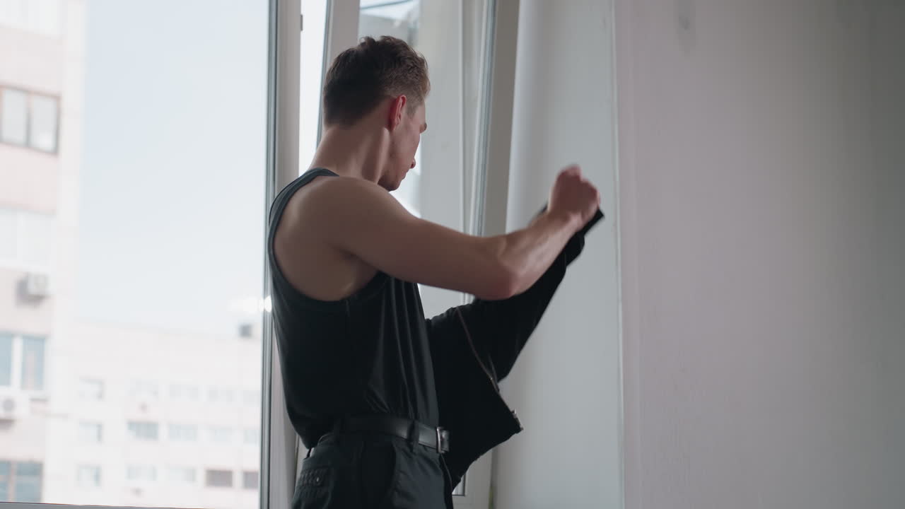 Young man in black tank top takes off black jacket while standing near large window in bright modern interior, soft natural light entering room, high rise city building visible through glass