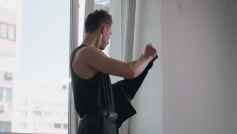 young man in black tank top takes off black jacket while standing near large window in bright modern interior, soft natural light entering room, high rise city building visible through glass