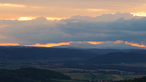 Hilly-landscape-lying-below-hills-full-of-farms-and-fields-during-sunset-with-orange-clouds